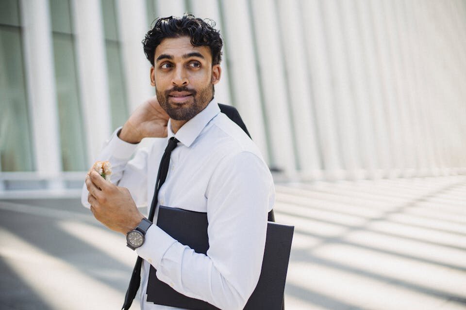 Businessman in formal attire eating a sandwich outdoors with a modern background.